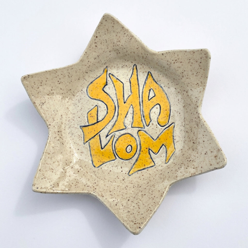 A six-pointed star-shaped ceramic dish with a speckled beige glaze, featuring the word 'SHA'LOM' in bold yellow graffiti-style lettering outlined in blue in the center.