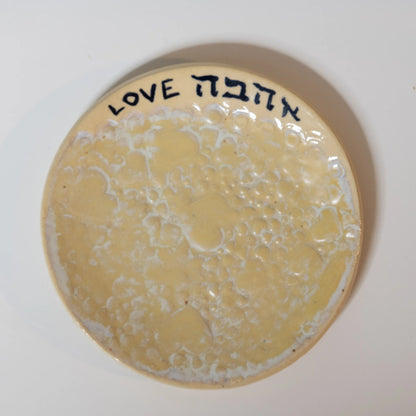 A round handmade ceramic plate with a textured, bubbled cream and light yellow glaze, featuring the word 'LOVE' in English and Hebrew ('אהבה') in black lettering along the rim, set against a plain white background.