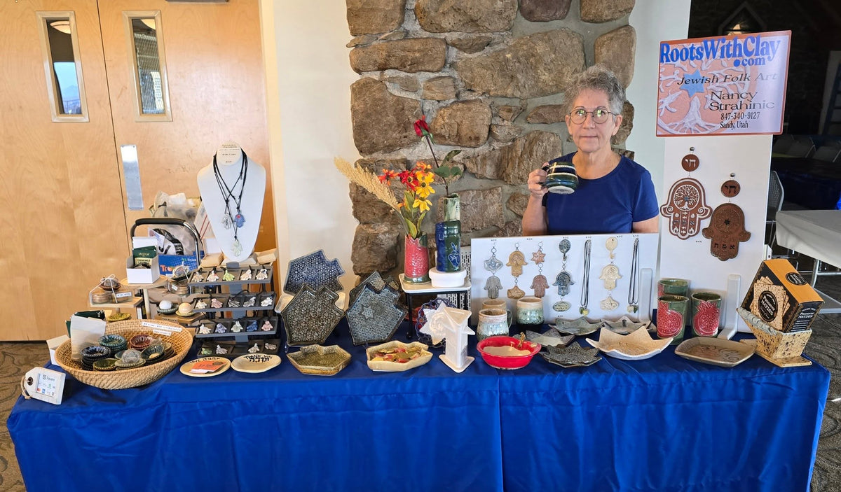 A craft fair booth display by Roots With Clay Studio, featuring a woman holding a mug behind a table covered in handmade jewelry, wooden items, and small craft supplies. A stone wall and wooden doors are visible in the background.