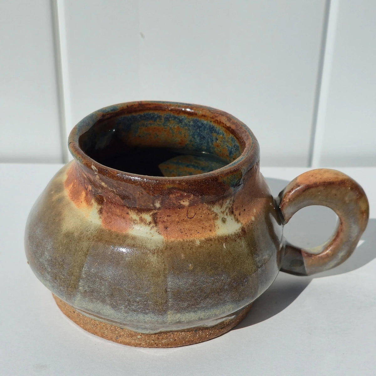 Angled view of a rounded stoneware belly mug showing its blue-green interior, rust-brown rim and circular handle