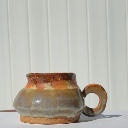 Side view of a rounded stoneware belly mug with gray-green glaze, a rust-brown rim, exposed clay base and loop handle